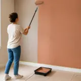 Mujer pintando una pared de color terracota con rodillo. Aprende como pintar paredes y transforma tu hogar.