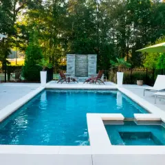Piscina moderna con jacuzzi y cascada de piedra, lista para el mantenimiento de piscinas. Sillas, sombrilla y vegetación completan el oasis.