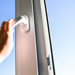 Mano girando la manija para reparar cierre ventana aluminio abatible. Cielo azul de fondo.