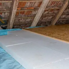 Aislamiento de suelo en una casa antigua con paneles de poliestireno sobre plástico azul, bajo vigas de madera y tejas.