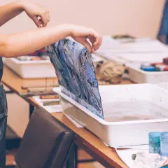 Manos sostienen tela con diseño marmoleado sobre una bandeja con agua, lista para la pintura para mármol.