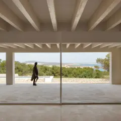 Persona camina por una terraza moderna con grandes ventanales que muestran varios tipos de ventanas correderas, vistas al mar y vegetación.