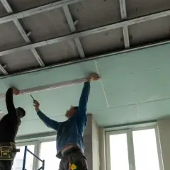 Dos obreros instalan placas de yeso para el aislamiento de techos. La estructura metálica del techo es visible.