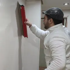 Hombre con gorra y chaqueta blanca alisa pared con llana, mostrando cómo alisar paredes de gotele para un acabado perfecto.