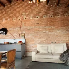 Cocina y sala de estar con paredes de ladrillo visto, un sofá beige y sillas de cuero. El acabado rústico en paredes aporta calidez.