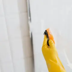 Mano con guante amarillo limpiando la mampara con esponja. Así se limpian las gomas de la mampara para un brillo impecable.