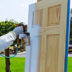 Persona con guantes y traje protector aplica pintura blanca a una puerta de madera con spray. Proceso de como lacar puertas barnizadas.