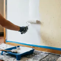 Persona aplicando pintura blanca con rodillo en pared, mostrando cómo hacer gotele con rodillo. Bandeja con pintura y brocha en el suelo.