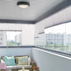 Balcón moderno con sofá, cojines coloridos y plantas. Las cortinas plisadas ofrecen privacidad, ideales para tus ideas para cubrir balcones.