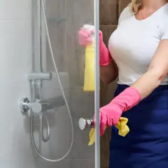 Persona con guantes rosas y delantal azul limpia la mampara del baño con spray amarillo.