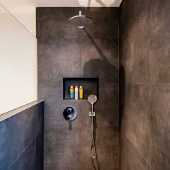 Duchas con murete y cristal. Cabezal de ducha, grifería y nicho con botes de champú. Azulejos oscuros y pared blanca.