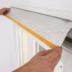 Manos colocando material aislante plateado con cinta amarilla para aislar cajón persiana.