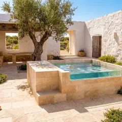 Piscina de obra pequeña con paredes de cristal, rodeada de piedra y un olivo. Ideal para aprender como hacer una piscina de obra pequeña.