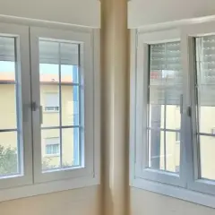 Dos ventanas batientes horizontales blancas con persianas enrollables. A través de ellas se ve un edificio amarillo con ventanas.