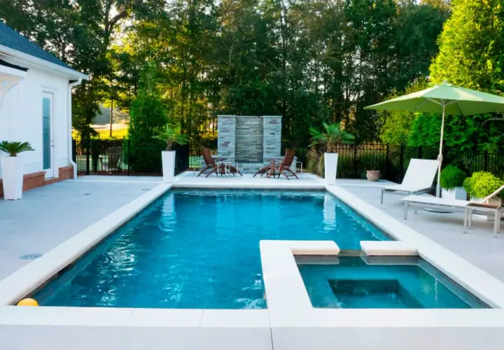 Piscina moderna con jacuzzi y cascada de piedra, lista para el mantenimiento de piscinas. Sillas, sombrilla y vegetación completan el oasis.