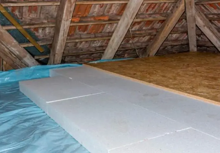 Aislamiento de suelo en una casa antigua con paneles de poliestireno sobre plástico azul, bajo vigas de madera y tejas.