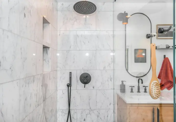 Ducha empotrada con grifo negro y rociador superior. La pared de mármol blanco tiene nichos empotrados.