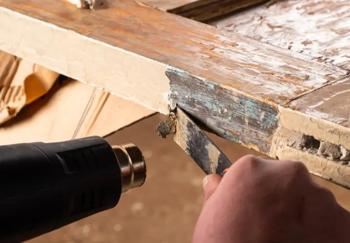Cómo quitar la pintura de la madera sin lijar: se usa pistola de calor y rasqueta para despegar capas viejas de pintura de un marco de madera.
