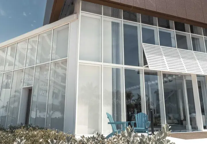 Edificio moderno con un **muro cortina fachada** de cristal. Dos sillas azules esperan en la entrada, reflejando palmeras.