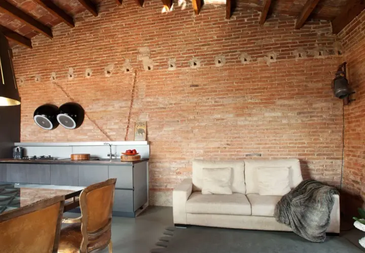 Cocina y sala de estar con paredes de ladrillo visto, un sofá beige y sillas de cuero. El acabado rústico en paredes aporta calidez.