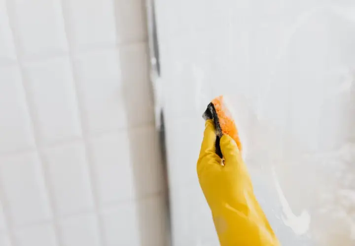 Mano con guante amarillo limpiando la mampara con esponja. Así se limpian las gomas de la mampara para un brillo impecable.