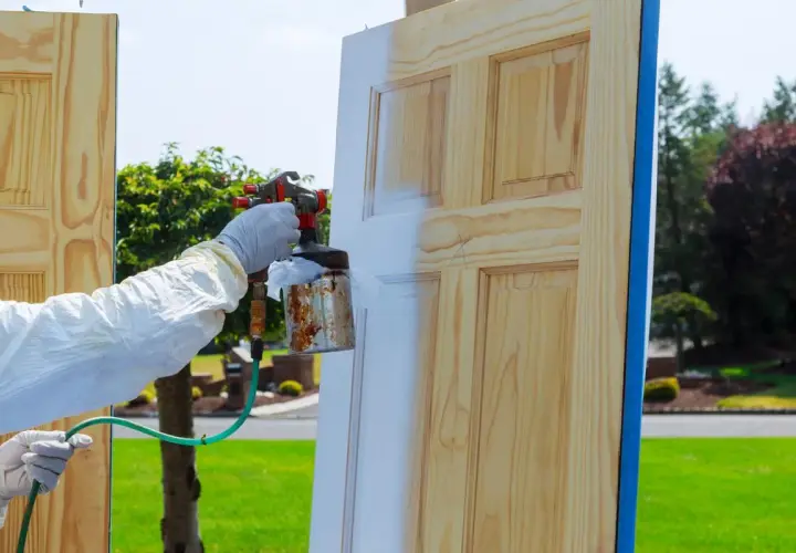 Persona con guantes y traje protector aplica pintura blanca a una puerta de madera con spray. Proceso de como lacar puertas barnizadas.
