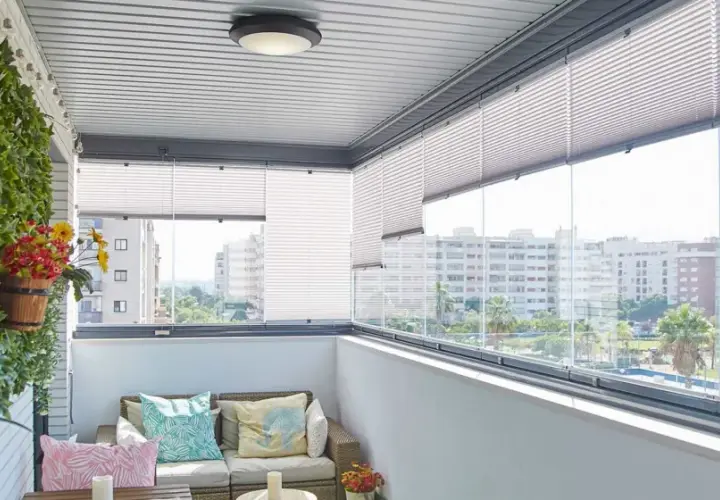 Balcón moderno con sofá, cojines coloridos y plantas. Las cortinas plisadas ofrecen privacidad, ideales para tus ideas para cubrir balcones.
