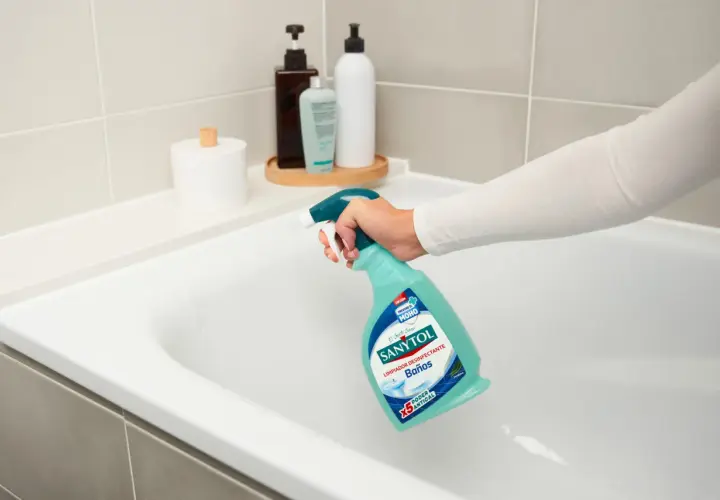 Mano rociando limpiador Sanytol en bañera blanca para quitar manchas oscuras plato ducha.
