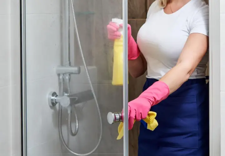 Persona con guantes rosas y delantal azul limpia la mampara del baño con spray amarillo.