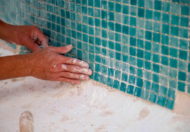 Manos trabajando en el cemento de piscinas elevadas de obra, colocando teselas turquesas.