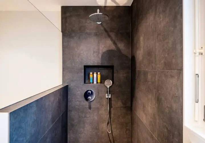 Duchas con murete y cristal. Cabezal de ducha, grifería y nicho con botes de champú. Azulejos oscuros y pared blanca.