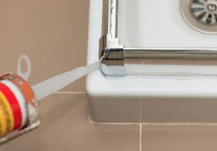 Aplicando sellador para mampara de ducha. Un aplicador de silicona deposita un cordón blanco para sellar la unión entre la mampara y la bañera.