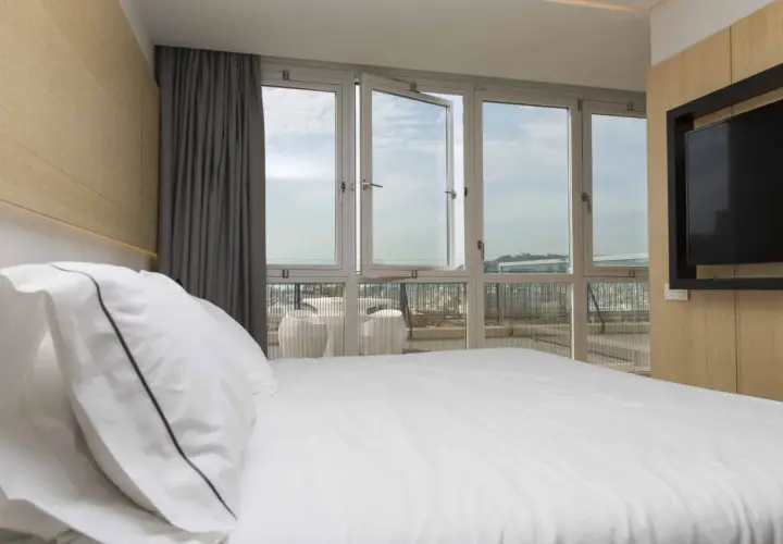 Cama blanca con almohadas, cortinas grises y ventanas de triple cristal que dejan ver el cielo y la ciudad.