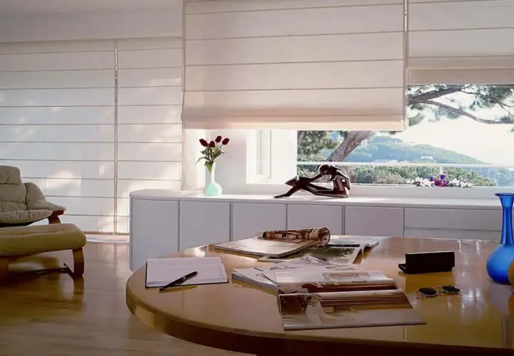 Estudio moderno con sillón, mesa y varios tipos de persianas romanas que filtran la luz del sol.