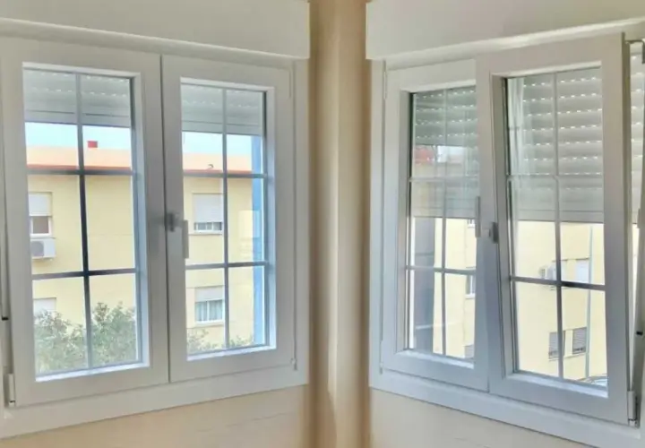 Dos ventanas batientes horizontales blancas con persianas enrollables. A través de ellas se ve un edificio amarillo con ventanas.