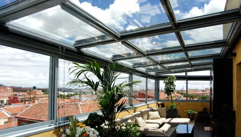 Disfruta de vistas panorámicas desde este cerramiento parcial terraza ático. Sofá cómodo, plantas y un cielo azul con nubes.