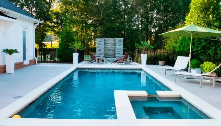Piscina moderna con jacuzzi y cascada de piedra, lista para el mantenimiento de piscinas. Sillas, sombrilla y vegetación completan el oasis.