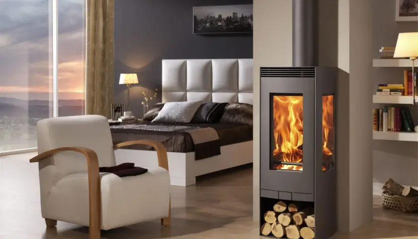 Estufa de leña moderna con fuego encendido, junto a una cama y sillón. Diversos tipos de estufas de gas ofrecen alternativas eficientes.