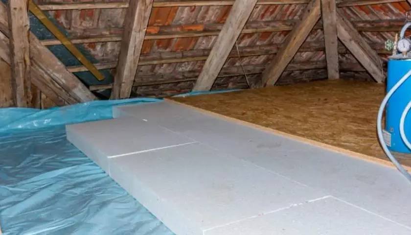 Aislamiento de suelo en una casa antigua con paneles de poliestireno sobre plástico azul, bajo vigas de madera y tejas.