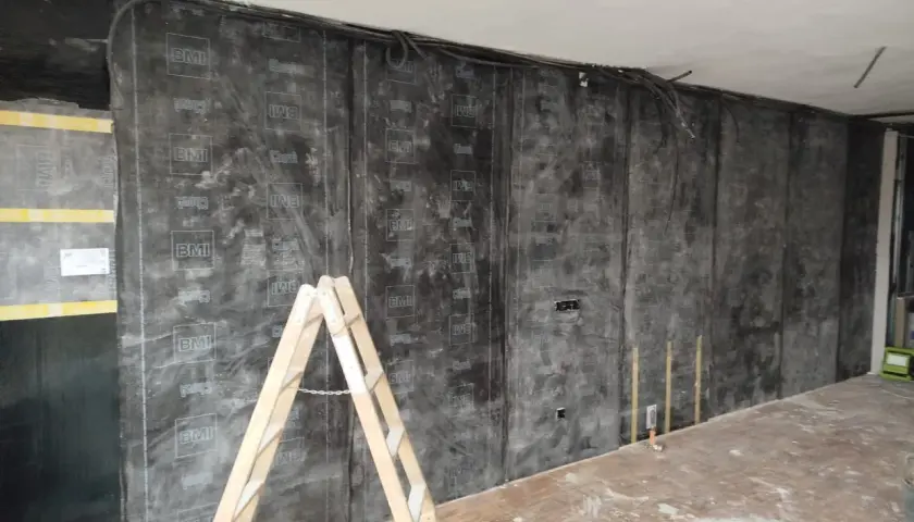 Instalación de material para insonorizar pared sin obra. Una escalera de madera apoya contra la pared cubierta con láminas negras.