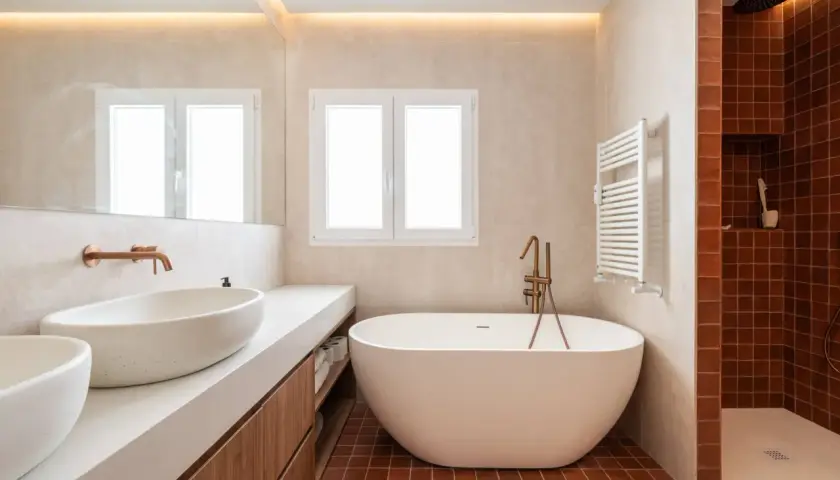 Baño moderno con bañera exenta y ducha. La iluminación indirecta en el techo y la luz natural de la ventana ayudan a como iluminar un baño.