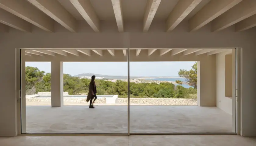 Persona camina por una terraza moderna con grandes ventanales que muestran varios tipos de ventanas correderas, vistas al mar y vegetación.