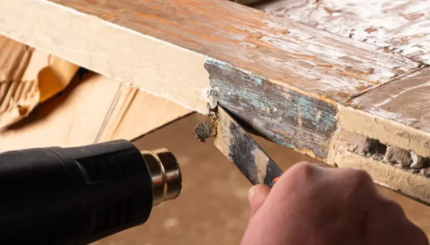 Cómo quitar la pintura de la madera sin lijar: se usa pistola de calor y rasqueta para despegar capas viejas de pintura de un marco de madera.