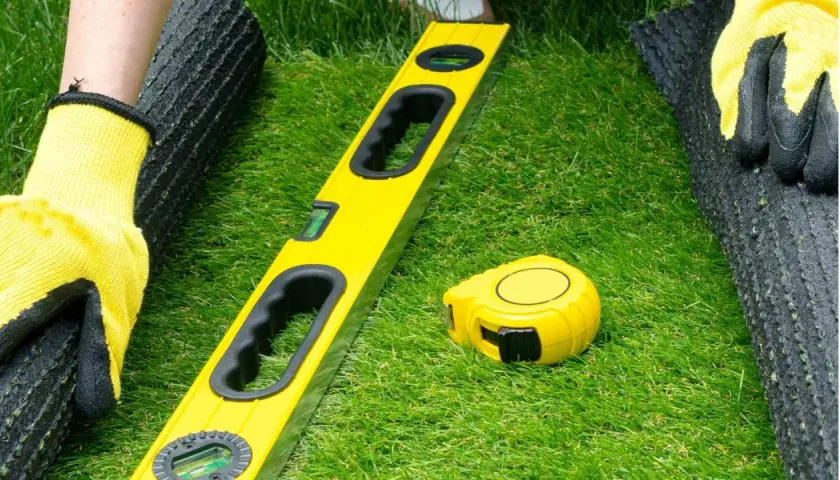 Manos enguantadas preparan el césped artificial. Nivel y cinta métrica listos para pegar.