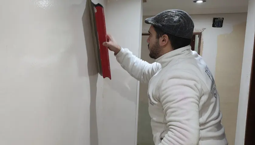 Hombre con gorra y chaqueta blanca alisa pared con llana, mostrando cómo alisar paredes de gotele para un acabado perfecto.