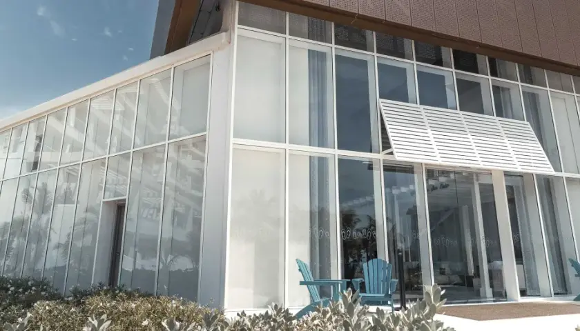 Edificio moderno con un **muro cortina fachada** de cristal. Dos sillas azules esperan en la entrada, reflejando palmeras.