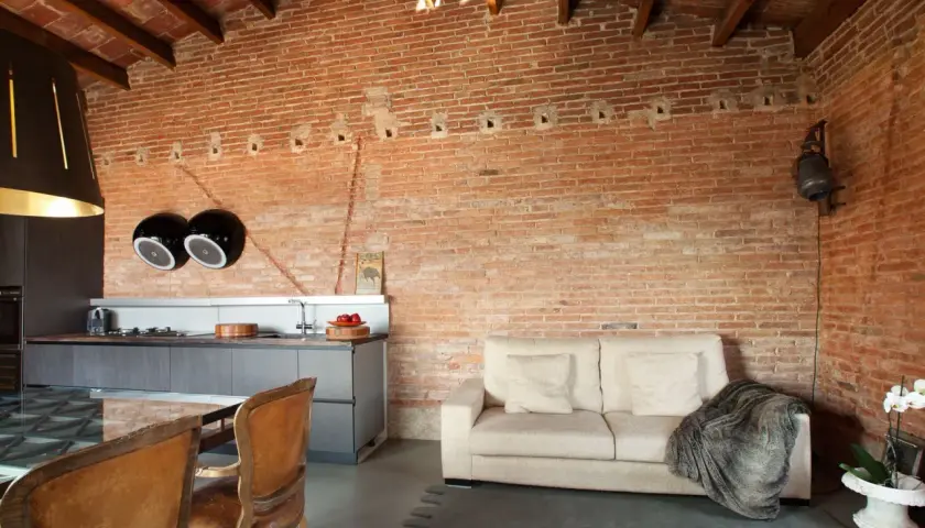 Cocina y sala de estar con paredes de ladrillo visto, un sofá beige y sillas de cuero. El acabado rústico en paredes aporta calidez.
