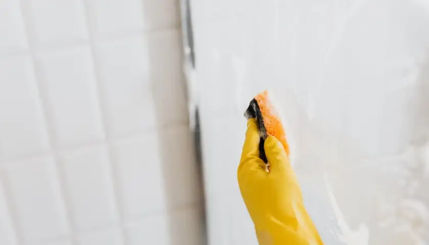 Mano con guante amarillo limpiando la mampara con esponja. Así se limpian las gomas de la mampara para un brillo impecable.