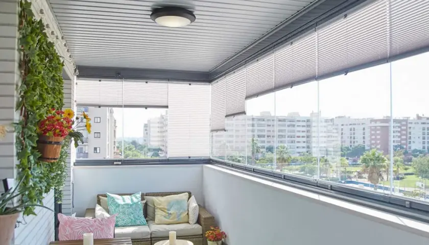 Balcón moderno con sofá, cojines coloridos y plantas. Las cortinas plisadas ofrecen privacidad, ideales para tus ideas para cubrir balcones.