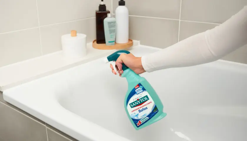 Mano rociando limpiador Sanytol en bañera blanca para quitar manchas oscuras plato ducha.